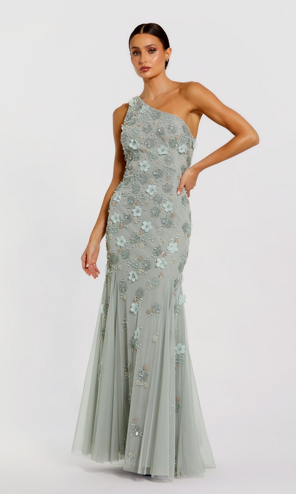A woman stands against a plain light background wearing the Long Formal Dress 9168 by Mac Duggal, featuring a light blue one-shoulder design with floral appliqués and delicate bead embellishments.